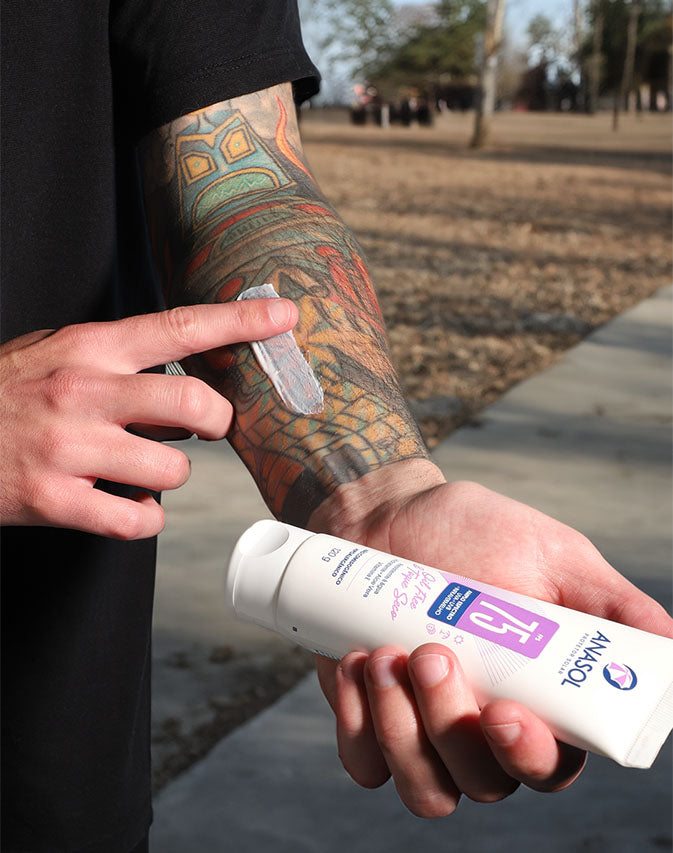 Homem espalhando o creme do corporal FPS 75 no antebraço tatuado, num parque, com a bisnaga na outra mão.