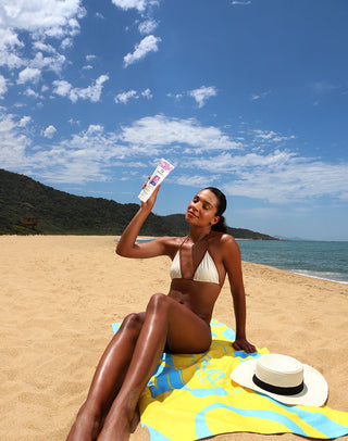 Mulher sentada na praia segurando o Protetor Solar Corporal Anasol FPS 75 sob o sol
