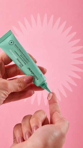 Mãos dispensando uma gota do Gel Secativo Antiacne Hidrabene na ponta do dedo para aplicação pontual, fundo rosa