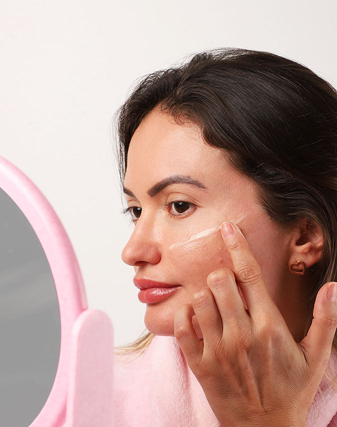 Mulher aplicando o creme Retinol Noturno na bochecha, diante do espelho.