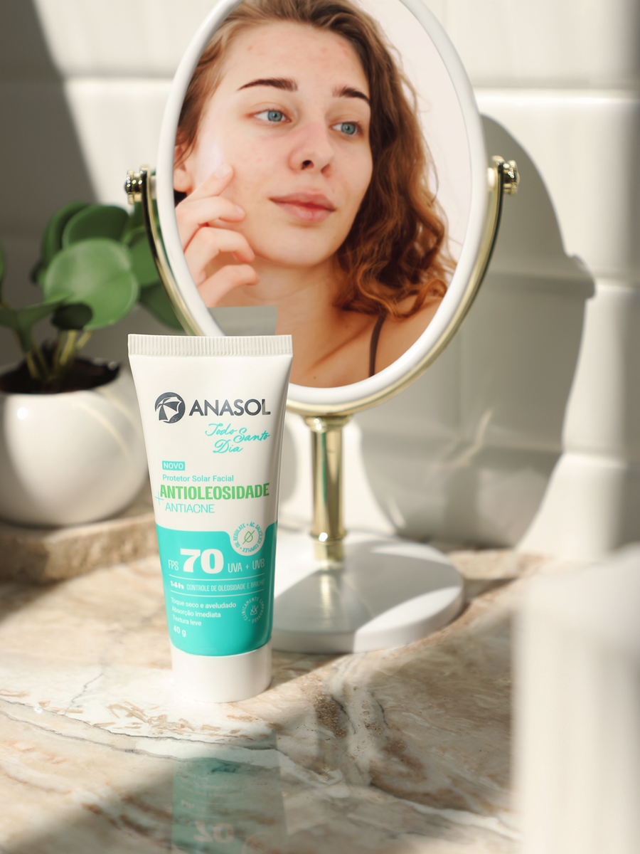 A bisnaga do Antiacne FPS 70 na bancada do banheiro, com uma mulher se olhando no espelho ao fundo.