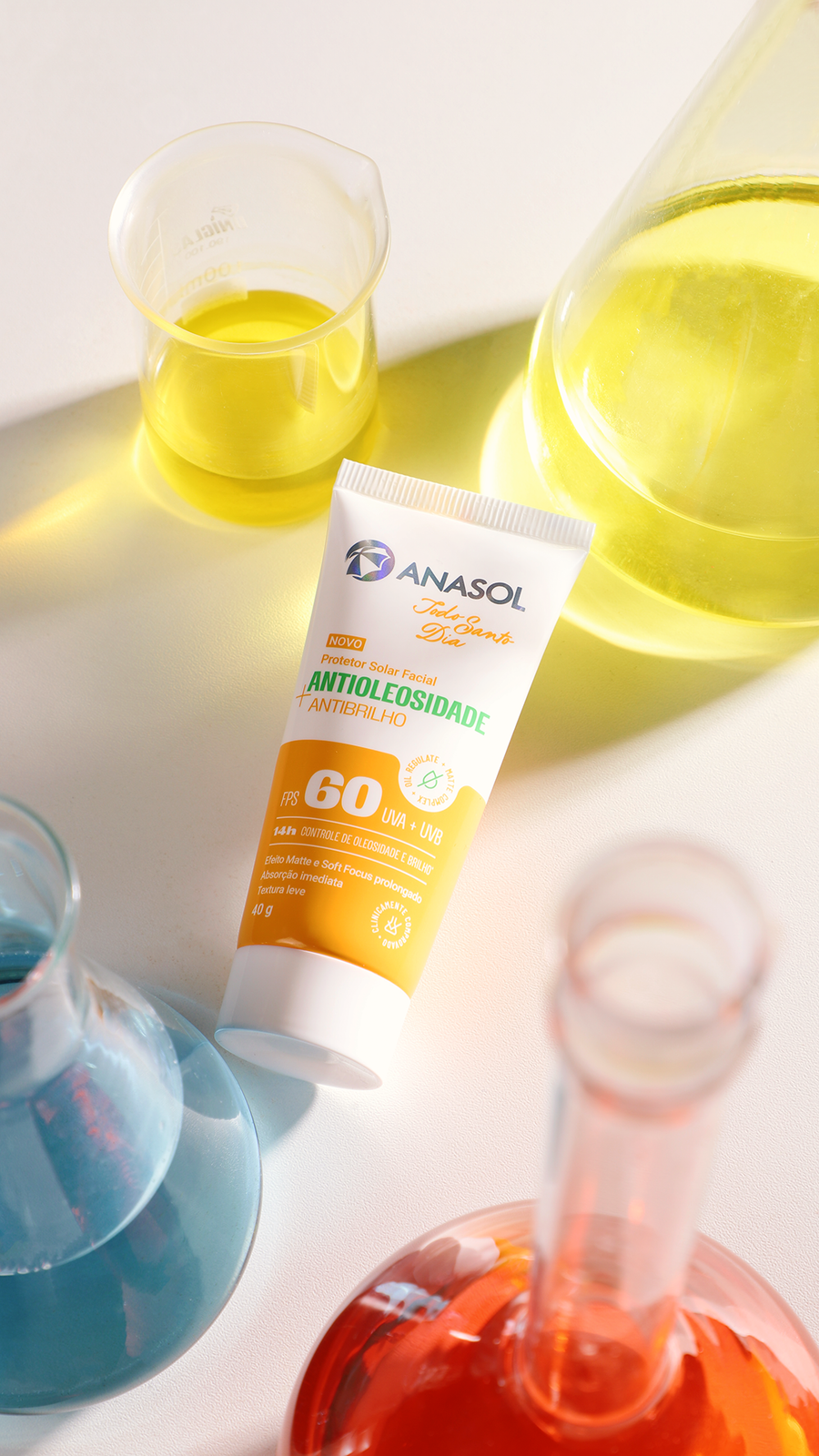 A bisnaga do Antibrilho FPS 60 deitada numa bancada clara, entre béqueres com líquidos amarelo, azul e laranja.