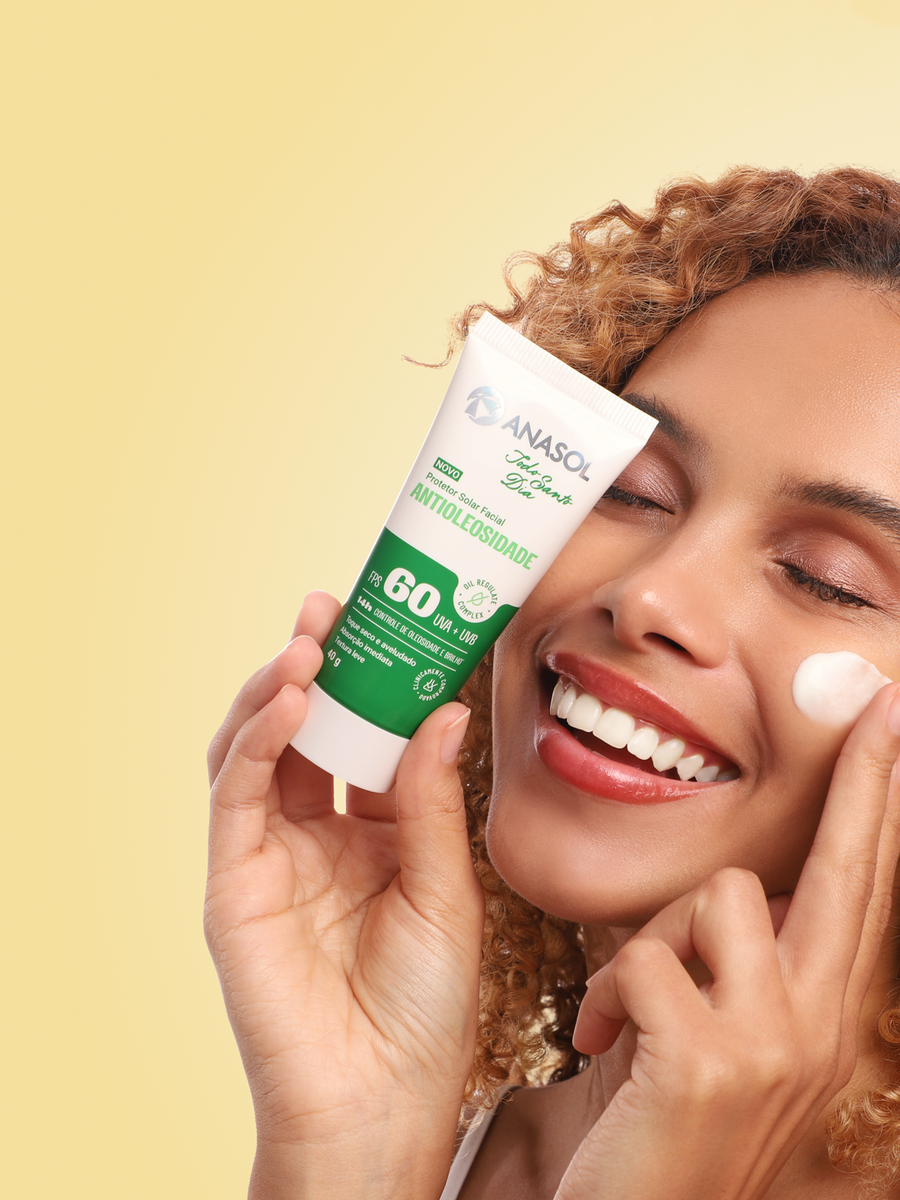 Mulher de cabelo cacheado sorrindo com um ponto de creme na bochecha, segurando a bisnaga do Antioleosidade FPS 60.