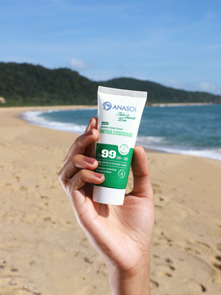 Mão segurando o protetor solar Anasol Todo Santo Dia FPS 99 na praia