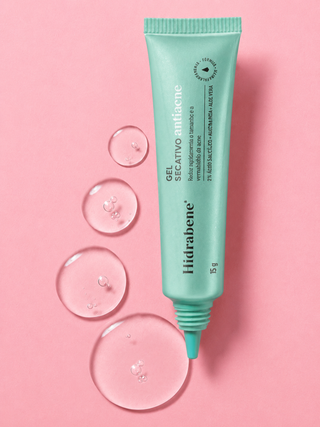 Bisnaga do Gel Secativo Antiacne Hidrabene deitada em fundo rosa entre gotas de gel